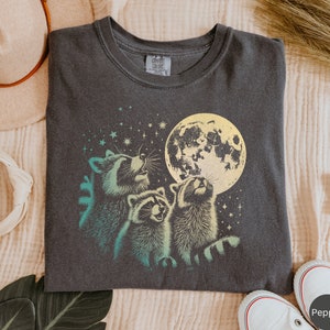 Op de afbeelding: Een donkergrijs T-shirt met een afbeelding van drie wasberen die naar een gele maan met sterren op de achtergrond kijken. De tekst "Comfort Colors" staat op het label.