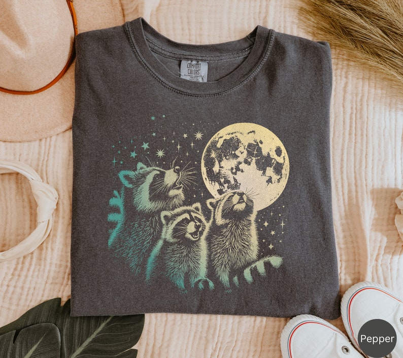 Op de afbeelding: Een donkergrijs T-shirt met een afbeelding van drie wasberen die naar een gele maan met sterren op de achtergrond kijken. De tekst "Comfort Colors" staat op het label.