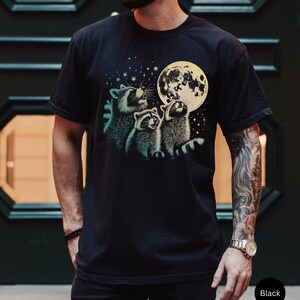 Op de afbeelding: Een zwart T-shirt met een grafische print van drie wasberen die naar een volle maan kijken met sterren op de achtergrond.