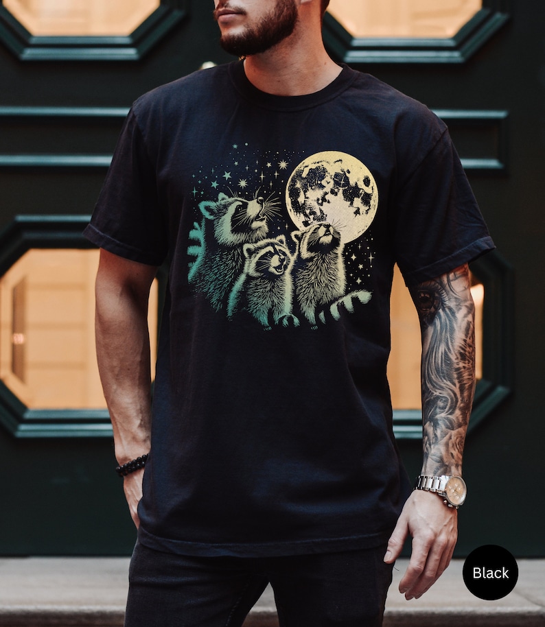 Op de afbeelding: Een zwart T-shirt met een grafische print van drie wasberen die naar een volle maan kijken met sterren op de achtergrond.