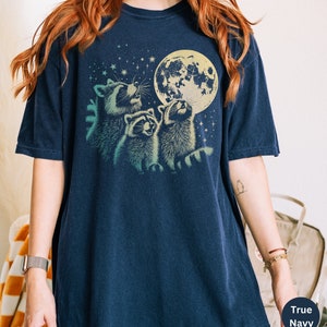 Op de afbeelding: Een marineblauw t-shirt met een afbeelding van drie wasberen die naar een volle maan met sterren op de achtergrond kijken.