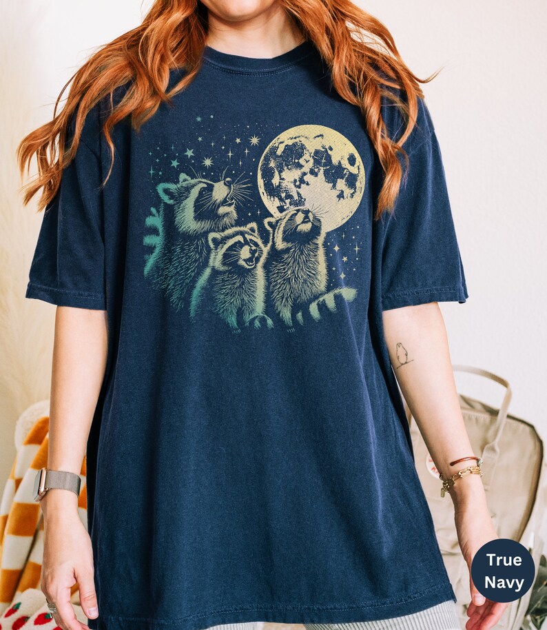 Op de afbeelding: Een marineblauw t-shirt met een afbeelding van drie wasberen die naar een volle maan met sterren op de achtergrond kijken.