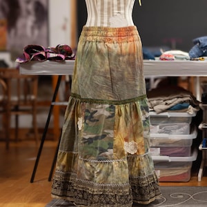 Può includere: Una lunga gonna patchwork su un manichino. La gonna presenta un motivo mimetico, inserti in pizzo e un design a balze. La vita è elasticizzata e i colori includono verde, marrone e arancione.
