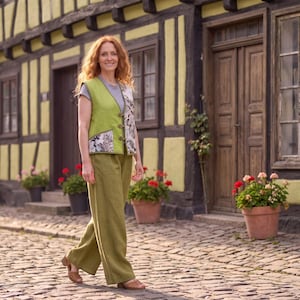May include: A lime green vest with a floral patterned panel and toggle closures, worn over a gray t-shirt, paired with wide-leg olive green trousers and brown sandals. The outfit features a mix of solid colors and botanical prints in a casual style.