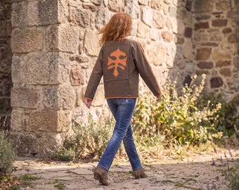 May include: A brown long-sleeve jacket featuring a large orange leaf-shaped applique on the back, orange trim along the sleeves and hem, paired with blue denim jeans and brown ankle boots with fringe detailing on the back of the heels.