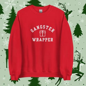 Könnte beinhalten: Ein rotes Sweatshirt mit dem weißen Text "GANGSTER WRAPPER" und einer Geschenkbox-Grafik. Das Sweatshirt besteht aus einem weichen Material. Der Hintergrund hat ein festliches Muster mit Schneeflocken, Weihnachtsbäumen und Rentieren.