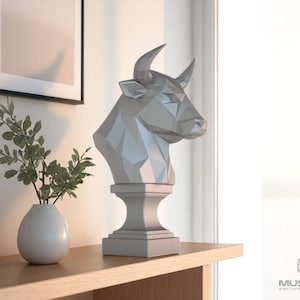 May include: A gray geometric bull bust sculpture on a pedestal, displayed on a wooden shelf. A white vase with green leaves is to the left. The bust is near a window, and a framed picture hangs on the wall.