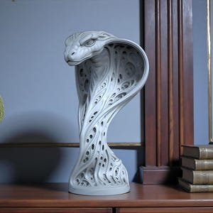 May include: A white cobra sculpture with an intricate, open design. The snake statue is displayed on a wooden surface. The background includes a wall, a mirror, and books. The sculpture's hood is open, showcasing a detailed pattern.