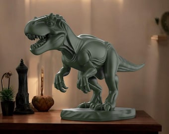 May include: A dark green Tyrannosaurus Rex sculpture on a rectangular base. The dinosaur has an open mouth, visible teeth, and detailed skin texture. The sculpture is displayed on a wooden surface with other decorative items.
