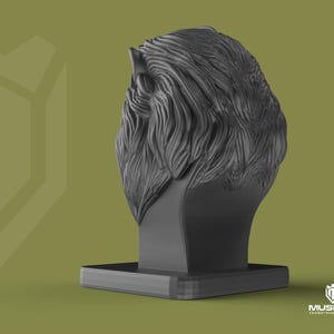May include: A dark grey sculpture of a lion's head with detailed fur texture, mounted on a rectangular base. The background is a muted olive green. The logo "MUSDAN INDUSTRIAL TASARIM" is visible in the bottom right corner.