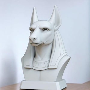 May include: A white bust of Anubis, the ancient Egyptian god of the afterlife, with a jackal head and human body. The sculpture features detailed facial features, tall ears, and a decorative collar. The bust is mounted on a rectangular base.