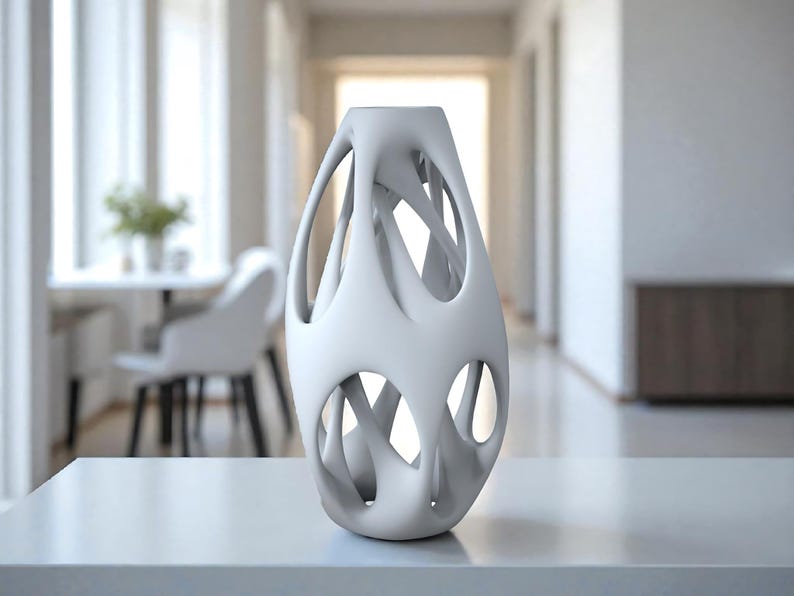 May include: A white, abstract ceramic vase with an open, lattice-like design. The vase has an elongated, oval shape with multiple openings and a smooth, matte finish. It is displayed on a white surface, suggesting a modern home decor item.