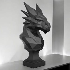 Może przedstawiać: Czarny, geometryczny gipsowy odlew smoka. Głowa smoka ma ostre, kanciaste detale i jest zamontowana na podstawie. Rzeźba jest wystawiona na szarej powierzchni na tle białej ściany.