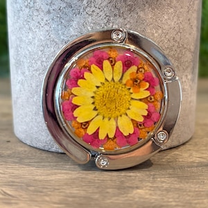 May include: A circular silver-toned metal bag hanger featuring a resin-encased floral arrangement. The center displays a large yellow daisy surrounded by a ring of small pink and orange dried flowers, framed by a polished metal border with rivets.