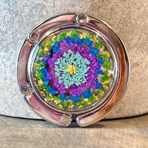 May include: A silver-toned purse hook with a circular, clear resin top. The resin contains a colorful floral arrangement of blue, purple, yellow, and green dried flowers. The hook is designed to hang from a table edge.