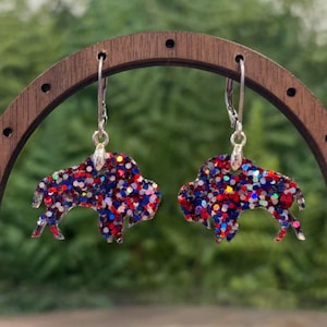 May include: A pair of dangle earrings featuring buffalo-shaped charms filled with red, white, and blue glitter. The charms are suspended from silver-toned metal ear wires, creating a festive look with a multi-colored, sparkling finish.
