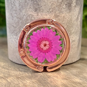 May include: A circular rose gold metal bag hanger featuring a pressed vibrant pink daisy flower encased in clear resin. The floral design is surrounded by small green foliage accents, and the metallic frame has a polished, reflective finish.
