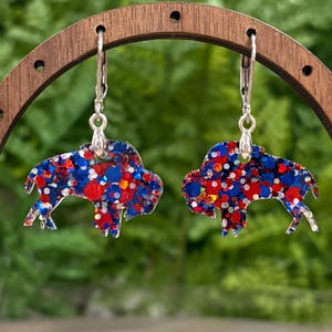 May include: A pair of buffalo-shaped earrings with lever-back closures. The earrings are filled with red, white, and blue glitter, creating a patriotic design. The earrings are displayed on a wooden stand against a blurred green background.