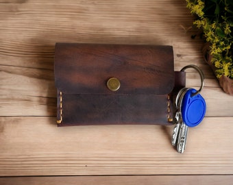 May include: A brown leather key holder with a gold button and a blue key fob attached to a silver key ring.