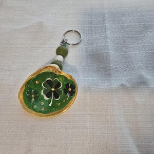 May include: A St. Patrick's Day themed keychain featuring a painted seashell. The shell is painted green with gold trim and a four-leaf clover design. The keychain has a silver ring and green, white, and olive-coloured beads.