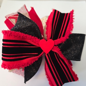 Puede incluir: Un lazo de rayas rojas y negras con un corazón rojo en el centro. El lazo tiene un borde deshilachado y está hecho de varias capas de tela.
