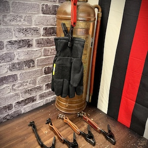 Puede incluir: Un juego de clips para guantes negros con una pinza de resorte de metal y una correa de cuero con cierre de botón a presión. Un clip está sujeto a un par de guantes de trabajo negros colgados de un extintor de cobre, otros cuatro están debajo.