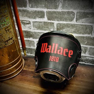 May include: A black leather cover featuring red stitching and the red text Wallace 1810 embossed on the front. The cover is shaped to fit over a circular object and includes black lacing details along the sides, providing a structured appearance.