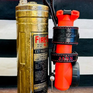 May include: A vintage brass fire extinguisher with a red and black label that reads "TRADE FIRE EXTINGUISHER TO OPEN TURN HANDLE PUMP STREAM", attached to a red and black leather keychain with the words "JANN" and "Nick Dillon Leatherworks" embossed on it.