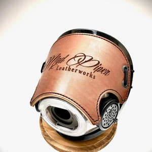 May include: A brown leather motorcycle helmet with a black base and a silver vent. The helmet has the text "Mad Piper Leatherworks" embossed on the front.