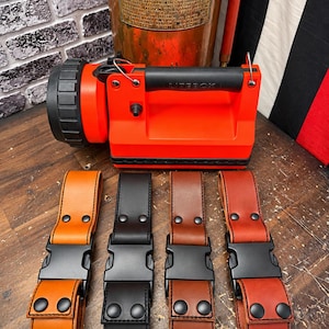 May include: A bright orange industrial lantern with a black handle and lens, positioned above four leather straps in tan, black, medium brown, and reddish-brown. Each strap features a black plastic side-release buckle and two black metal rivets.