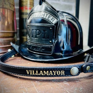 May include: A black leather firefighter helmet band featuring the name VILLAMAYOR embossed in gold lettering, attached to a black firefighter helmet with a front plate displaying the text FIREFIGHTER and MPL in raised black letters.