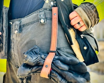 May include: Close-up of a firefighter's gear, featuring black leather gloves attached to a tan leather strap with the text "Mr. Fire". The strap is connected to the firefighter's gray pants and black jacket with reflective yellow stripes.