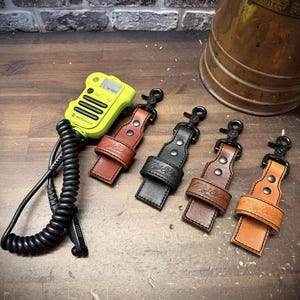 May include: A neon yellow Motorola radio with a coiled black cord and four leather radio straps in tan, black, dark brown, and light brown. Each strap features a metal swivel clip, two rivets, and an embossed logo on the front leather loop.