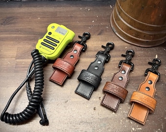 Op de afbeelding: Een neongele Motorola-radio met een zwart spiraalsnoer en vier leren radiohouders in lichtbruin, zwart, donkerbruin en beige. Elke houder is voorzien van een draaibare metalen clip, twee klinknagels en een in reliëf gedrukt logo op de lus.