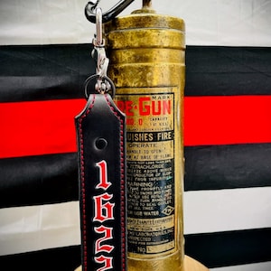 May include: A black leather keychain with red stitching and the number 1622 in red. The keychain is attached to a vintage brass fire extinguisher with the words "TRADE MARK RE-GUN" and "UISHES FIRE" printed on the side.