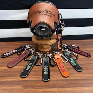 May include: A brown leather camera cover with the text Mad Piper Leatherworks, displayed on a wooden stand. Surrounding the stand are several leather keychains in black, brown, and orange, each featuring metal carabiners and personalized text.