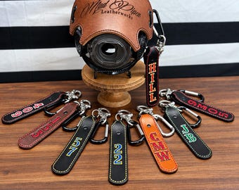 May include: A brown leather camera cover with the text Mad Piper Leatherworks, displayed on a wooden stand. Surrounding the stand are several leather keychains in black, brown, and orange, each featuring metal carabiners and personalized text.