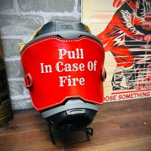 May include: A black MSA helmet featuring a red leather front piece with white stitched edges. The text "Pull In Case Of Fire" is printed in white lettering on the center of the red leather. The helmet is mounted on a black wire stand.