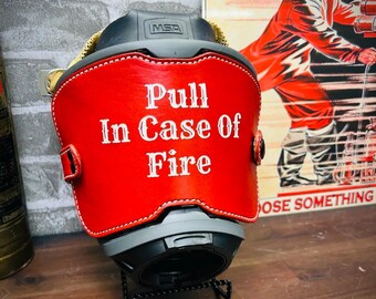 May include: A black MSA helmet featuring a red leather front piece with white stitched edges. The text "Pull In Case Of Fire" is printed in white lettering on the center of the red leather. The helmet is mounted on a black wire stand.