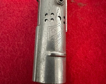 Peut inclure: Briquet en métal argenté avec un motif de flamme et le mot "BOWERS" gravé sur le côté. Le briquet est doté d'une petite chaîne attachée au bas.