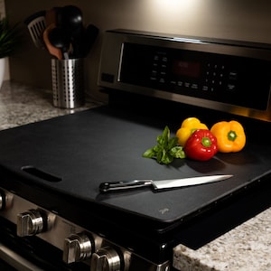 May include: A black silicone stovetop protector with a handle, placed on a black electric hob. A knife and a few bell peppers are on the protector.