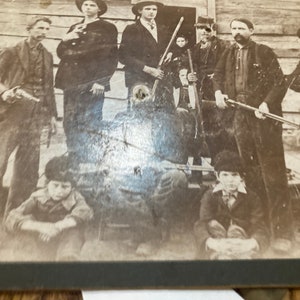 May include: A black and white vintage photograph of a group of men and boys posing with guns. The men are wearing hats and suits, and the boys are wearing jackets and pants. The group is standing in front of a wooden building.