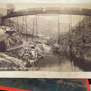 May include: A black and white photograph of a train crossing a high bridge over a river canyon. The text "HIGH BRIDGE ON THE LOOP GEORGETOWN COLO." is printed at the bottom of the image.