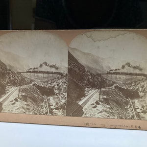 May include: A vintage stereoscopic photograph of a train traveling through a mountain pass. The train is on a trestle bridge and is emitting smoke. The image is captioned "Meadville, Pa. St. Louis, Mo. Copyright 1898, by B. L. Singlay."
