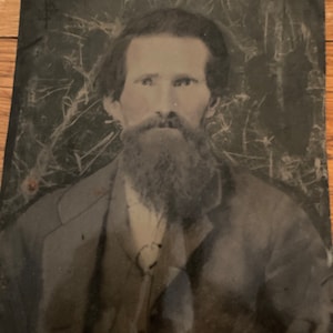 May include: A black and white portrait of a man with a long beard. He is wearing a dark jacket and a light-colored shirt. The image is faded and has some scratches.