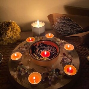 Pode incluir: Uma bandeja de madeira com uma tigela preta no centro, cercada por pequenas pedras coloridas e velas de ch&aacute; acesas. A tigela cont&eacute;m um l&iacute;quido preto e uma &uacute;nica vela de ch&aacute; acesa. Uma vela branca est&aacute; no fundo.