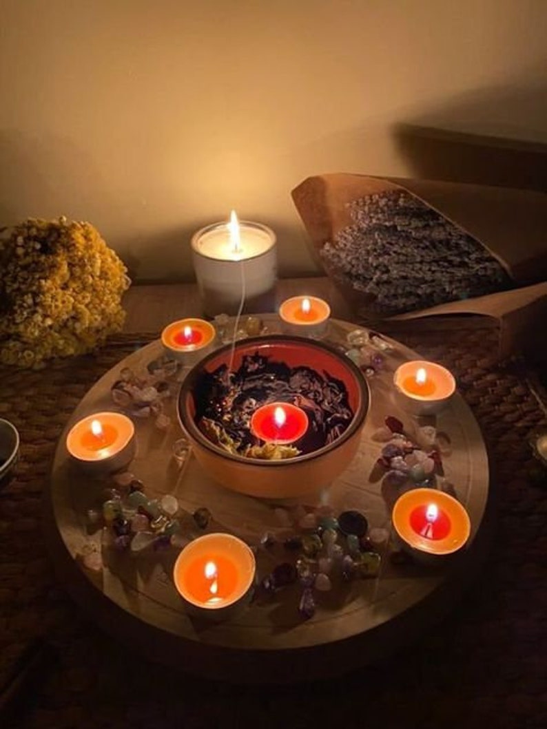 Pode incluir: Uma bandeja de madeira com uma tigela preta no centro, cercada por pequenas pedras coloridas e velas de ch&aacute; acesas. A tigela cont&eacute;m um l&iacute;quido preto e uma &uacute;nica vela de ch&aacute; acesa. Uma vela branca est&aacute; no fundo.