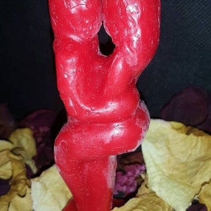Pode incluir: Uma vela vermelha em forma de duas figuras abra&ccedil;ando-se. A vela est&aacute; sobre uma base e tem uma superf&iacute;cie texturizada.