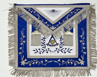 Puede incluir: Delantal masónico azul y blanco con flecos plateados y detalles bordados, incluyendo un compás y una escuadra, un ojo que todo lo ve y diseños florales.