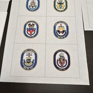 May include: A white fabric with six blue and gold embroidered patches of US Navy ship crests. The crests include the USS Wasp LHD 1, USS Green Bay LPD 20, USS Ashland LSD 48, USS Patriot MCM 7, USS Chief MCM 14, and USS Barry DDG 52.